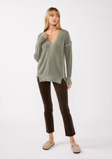 [Color: Sage/Natural] A blonde model wearing an everyday green sweater with a relaxed silhouette. With a classic v neckline, long sleeves, and an exposed seam with blanket stitch. A fall sweater styled with black bottom and casual sandals. 