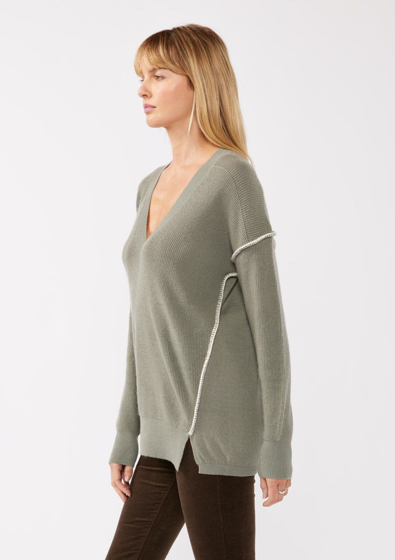 [Color: Sage/Natural] A blonde model wearing an everyday green sweater with a relaxed silhouette. With a classic v neckline, long sleeves, and an exposed seam with blanket stitch. A fall sweater styled with black bottom and casual sandals. 