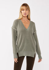 [Color: Sage/Natural] A blonde model wearing an everyday green sweater with a relaxed silhouette. With a classic v neckline, long sleeves, and an exposed seam with blanket stitch. A fall sweater styled with black bottom and casual sandals. 