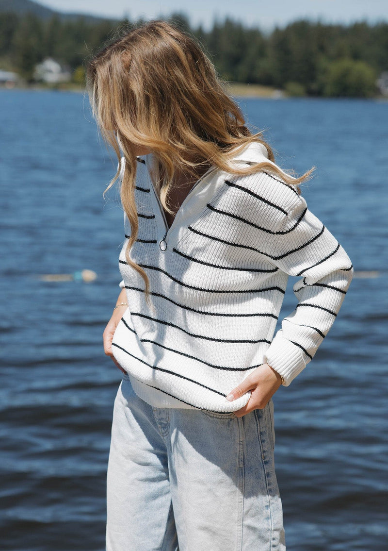 [Color: White/Black] A blonde model wearing a cozy knit stripe pullover sweater in black and white. With a zip-up funnel neck with v neckline, long sleeve, and ribbed hem and sleeves. A relaxed fit sweater styled with denim jeans for an ultra boho fall look.