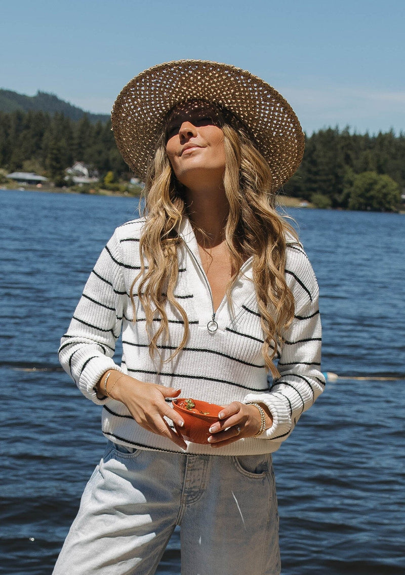 [Color: White/Black] A blonde model wearing a cozy knit stripe pullover sweater in black and white. With a zip-up funnel neck with v neckline, long sleeve, and ribbed hem and sleeves. A relaxed fit sweater styled with denim jeans for an ultra boho fall look.