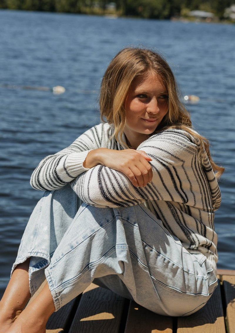 [Color: Natural/Black] A blonde model wearing a cozy striped sweater in ivory and black. A pullover sweater with long sleeves, v neckline, and ribbed cuffs and hem. Styled with relaxed denim for an ultra cozy summer to fall look.