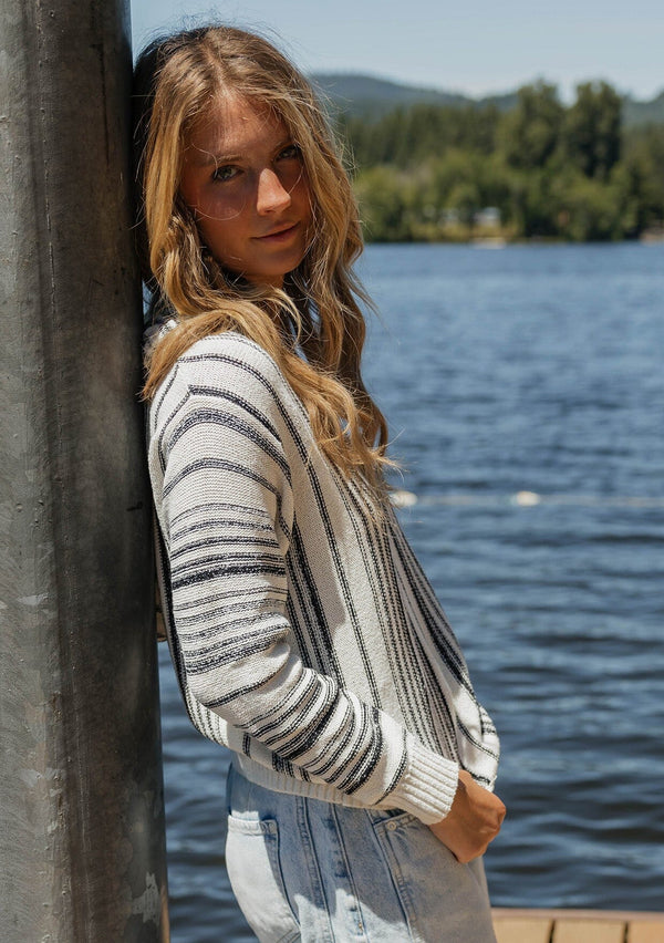 [Color: Natural/Black] A blonde model wearing a cozy striped sweater in ivory and black. A pullover sweater with long sleeves, v neckline, and ribbed cuffs and hem. Styled with relaxed denim for an ultra cozy summer to fall look.