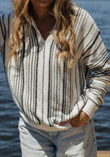 [Color: Natural/Black] A blonde model wearing a cozy striped sweater in ivory and black. A pullover sweater with long sleeves, v neckline, and ribbed cuffs and hem. Styled with relaxed denim for an ultra cozy summer to fall look.