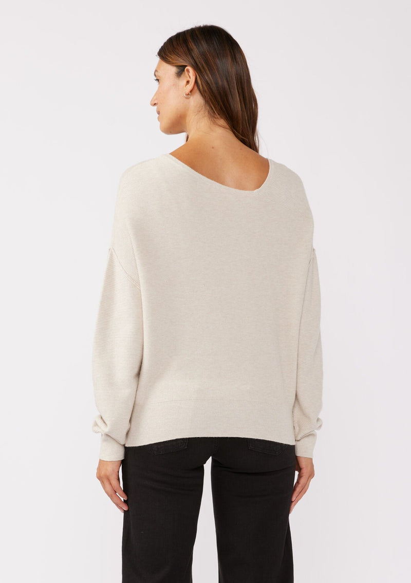 [Color: Heather Stone] A brunette model wearing a beige off white relaxed fit sweater with an asymmetrical neckline and long sleeves with a dropped shoulder. An everyday pullover sweater styled with dark denim jeans for the fall season.