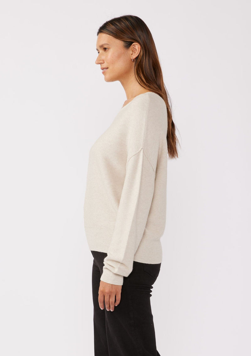 [Color: Heather Stone] A brunette model wearing a beige off white relaxed fit sweater with an asymmetrical neckline and long sleeves with a dropped shoulder. An everyday pullover sweater styled with dark denim jeans for the fall season.