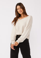 [Color: Heather Stone] A brunette model wearing a beige off white relaxed fit sweater with an asymmetrical neckline and long sleeves with a dropped shoulder. An everyday pullover sweater styled with dark denim jeans for the fall season.