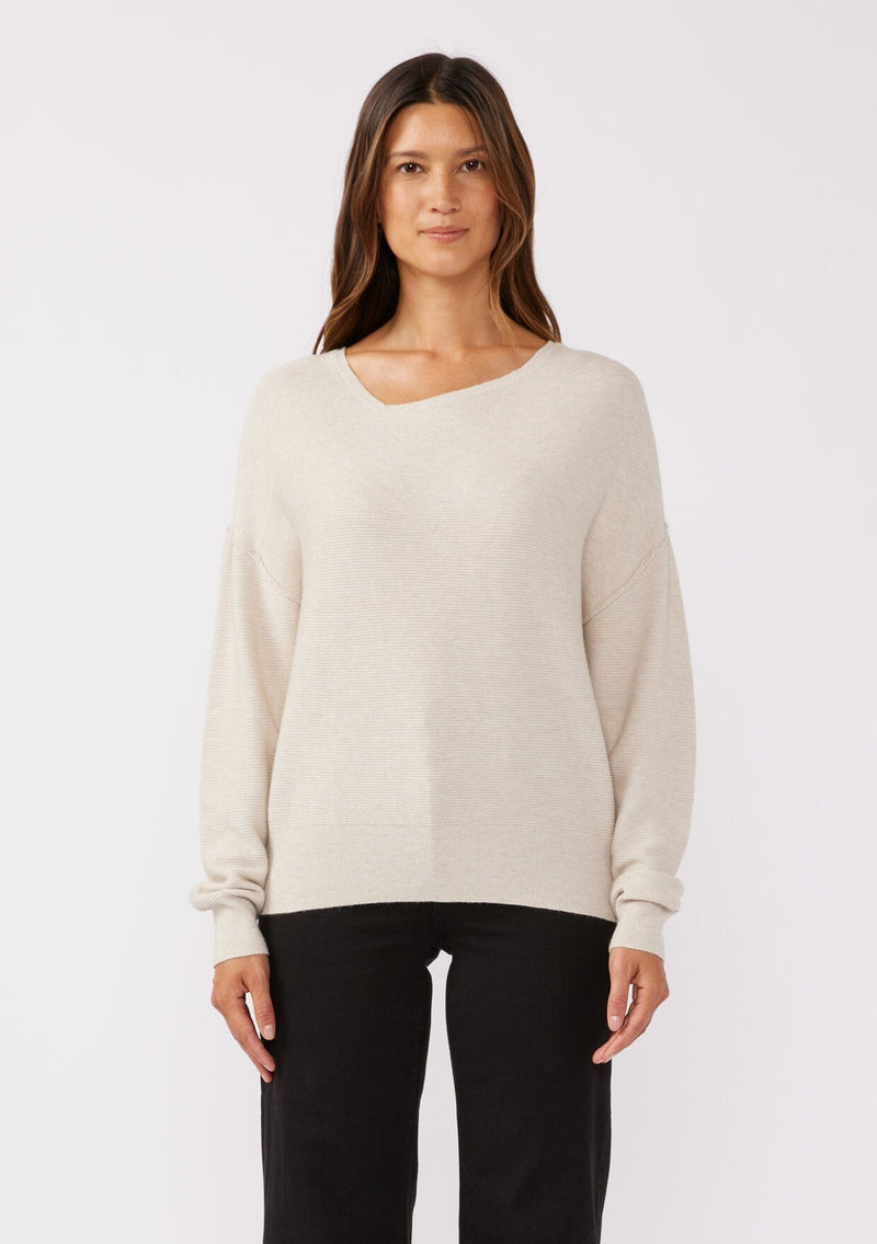 [Color: Heather Stone] A brunette model wearing a beige off white relaxed fit sweater with an asymmetrical neckline and long sleeves with a dropped shoulder. An everyday pullover sweater styled with dark denim jeans for the fall season.