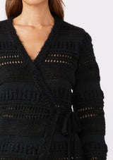 [Color: Black] A brunette model wearing a black sweater knit top with a wrap front and side tie closure. A chunky knit sweater with a mixed yarn weave and features an open knit design with a v neckline and long sleeves. 