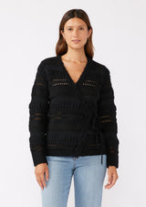 [Color: Black] A brunette model wearing a black sweater knit top with a wrap front and side tie closure. A chunky knit sweater with a mixed yarn weave and features an open knit design with a v neckline and long sleeves. 