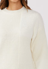 [Color: Cream] A detail shot of a brunette model wearing a fuzzy soft sweater crafted in a mixed media patchwork. With a crew neckline, long sleeves, and a relaxed fit. This cozy pullover sweater in cream ivory is the perfect addition to any Fall wardrobe. 