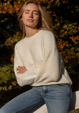 [Color: Cream] A lifestyle image of a blonde model wearing a fuzzy soft sweater crafted in a mixed media patchwork. With a crew neckline, long sleeves, and a relaxed fit. This cozy pullover sweater in cream ivory is the perfect addition to any Fall wardrobe. 