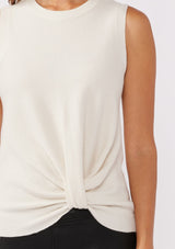 [Color: Vanilla] A detail shot of a brunette model wearing a cozy off white sweater tank top with a ribbed texture, twisted front hemline and a round neckline. A relaxed casual top for the spring season. 