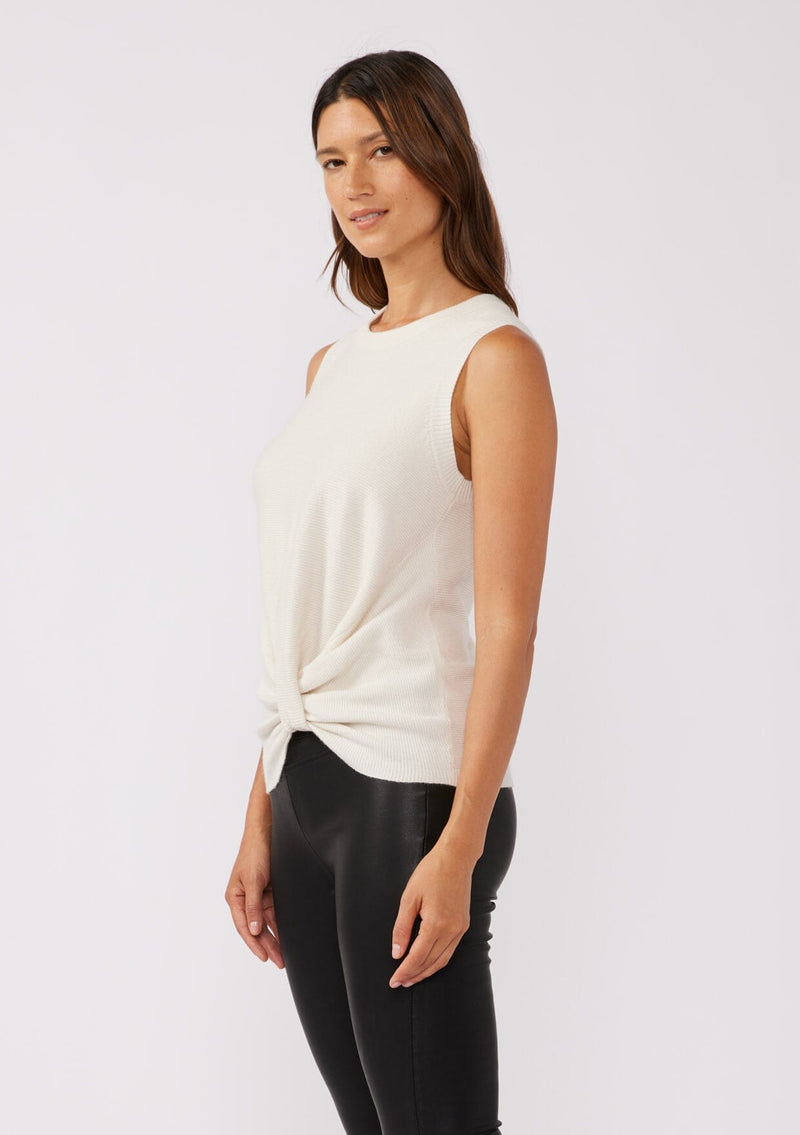 [Color: Vanilla] A side facing image of a brunette model wearing a cozy off white sweater tank top with a ribbed texture, twisted front hemline and a round neckline. A relaxed casual top for the spring season. 