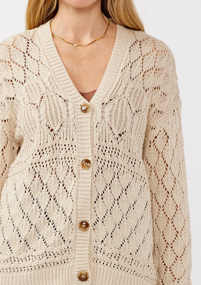 [Color: Vanilla] A detail image of a blonde model wearing wearing a light 
beige pointelle cardigan with an open knit design, button front, v-neckline, long sleeves, and ribbed trim, ideal for versatile spring layering.