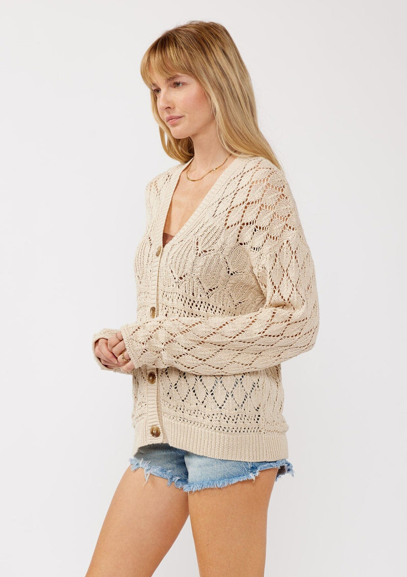 [Color: Vanilla] A side facing image of a blonde model wearing wearing a light 
beige pointelle cardigan with an open knit design, button front, v-neckline, long sleeves, and ribbed trim, ideal for versatile spring layering.