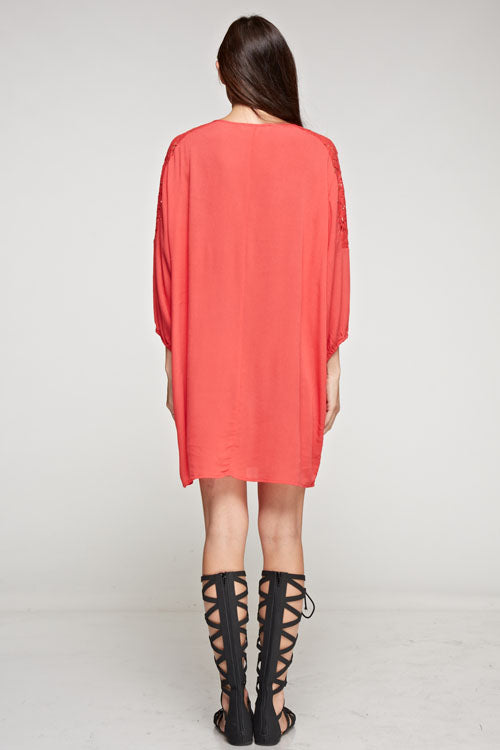 [Color: Poppy] A brunette model wearing a coral relaxed-fit mini dress with long sleeves, elastic cuffs, lace shoulder accents, and a split V-neckline with tie detail—a breezy beach cover-up.