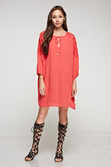 [Color: Poppy] A brunette model wearing a coral relaxed-fit mini dress with long sleeves, elastic cuffs, lace shoulder accents, and a split V-neckline with tie detail—a breezy beach cover-up.