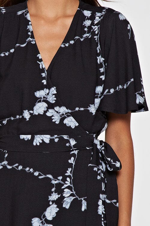 [Color: Black Combo] A detail image of a brunette model wearing a  black flowy wrap dress with a floral print. A boho dress with a short flutter sleeves, surplice v-neckline, and a side tie waist closure. 