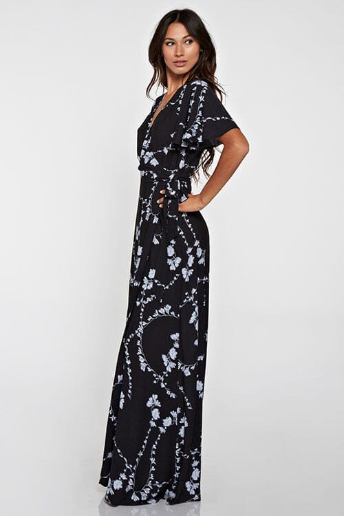 [Color: Black Combo] A side facing image of a brunette model wearing a  black flowy wrap dress with a floral print. A boho dress with a short flutter sleeves, surplice v-neckline, and a side tie waist closure. 