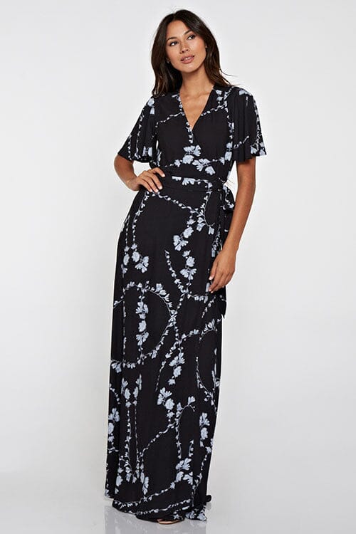 [Color: Black Combo] A front facing image of a brunette model wearing a  black flowy wrap dress with a floral print. A boho dress with a short flutter sleeves, surplice v-neckline, and a side tie waist closure. 