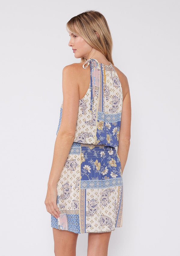 [Color: Ivory/Lilac] A back facing image of a blonde model wearing a cute summer mini dress designed in an ivory and lilac purple patchwork floral print. With a tie neckline, a front keyhole detail, and a smocked elastic waist.