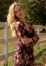 [Color: Plum/Dusty Rose] A detail lifestyle image of a blonde model wearing a sheer, empire waist maxi dress with a purple floral pattern. This flowy relaxed dress features a v-neckline, long sleeves, a button front, a ruffle cuff, and a tiered skirt. A fall special occasion dress for weddings, holiday events, and more. 