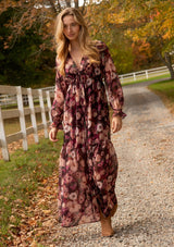 [Color: Plum/Dusty Rose] A full body front facing lifestyle image of a blonde model wearing a sheer, empire waist maxi dress with a purple floral pattern. This flowy relaxed dress features a v-neckline, long sleeves, a button front, a ruffle cuff, and a tiered skirt. A fall special occasion dress for weddings, holiday events, and more. 