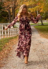 [Color: Plum/Dusty Rose] A full body front facing lifestyle image of a blonde model wearing a sheer, empire waist maxi dress with a purple floral pattern. This flowy relaxed dress features a v-neckline, long sleeves, a button front, a ruffle cuff, and a tiered skirt. A fall special occasion dress for weddings, holiday events, and more. 