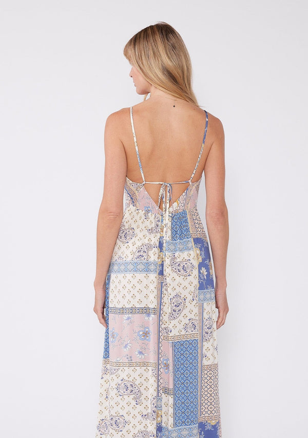 [Color: Ivory/Lilac] A back facing image of a blonde model wearing a sexy strappy maxi dress in a bohemian ivory and lilac purple patchwork floral print. With two side slits, a flattering v neckline, and adjustable spaghetti straps.
