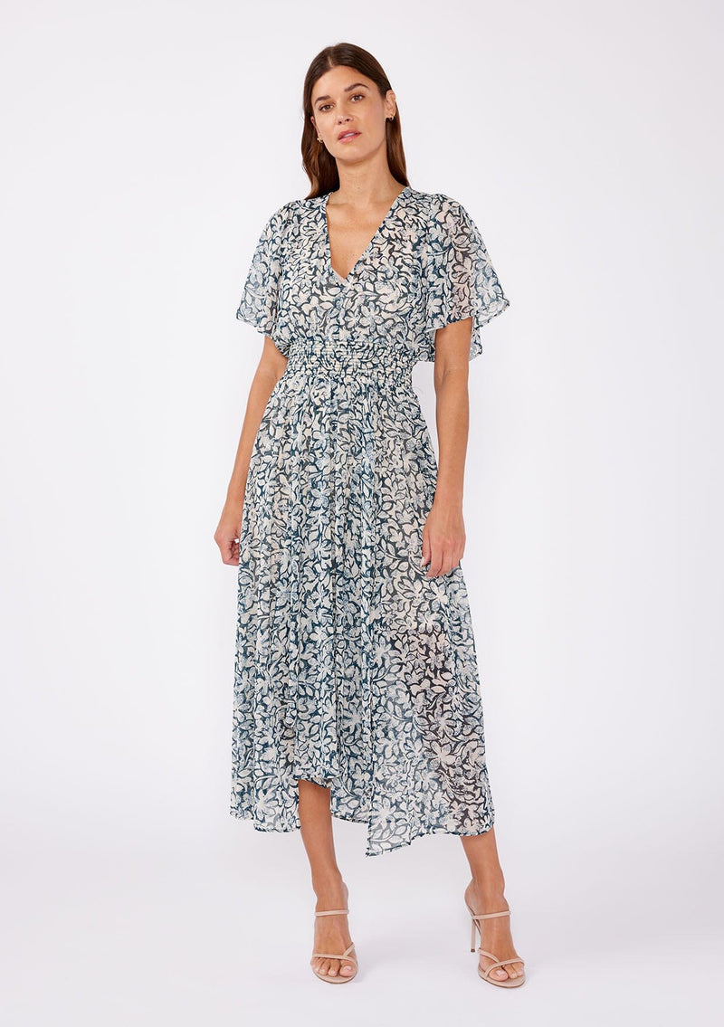 [Color: Teal/Taupe] A brunette woman wearing a summer to fall transitional midi dress with floral print on a chiffon like fabric. With a flattering v neckline, smocked waistline and an asymmetrical hem with multiple slits. 