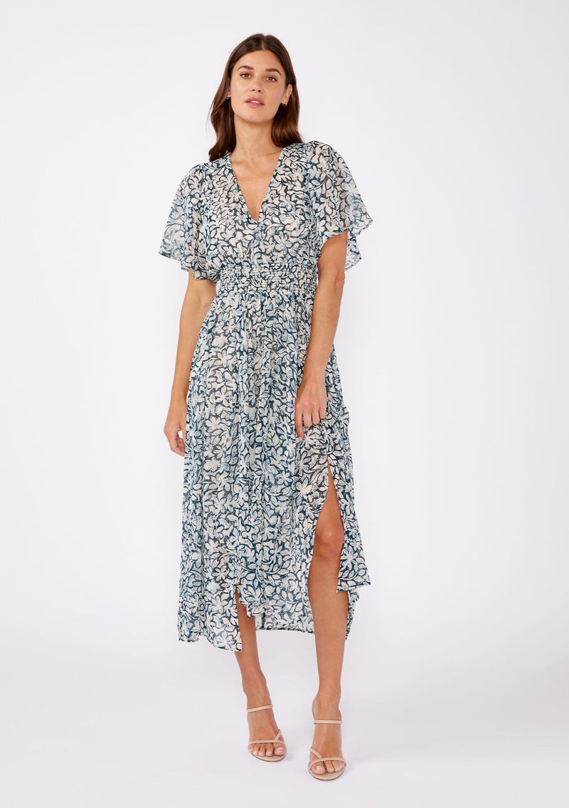 [Color: Teal/Taupe] A brunette woman wearing a summer to fall transitional midi dress with floral print on a chiffon like fabric. With a flattering v neckline, smocked waistline and an asymmetrical hem with multiple slits.