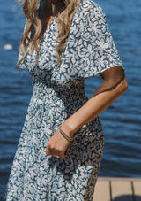 [Color: Teal/Taupe] A blonde woman wearing a summer to fall transitional midi dress with floral print on a chiffon like fabric. With a flattering v neckline, smocked waistline and an asymmetrical hem with multiple slits.