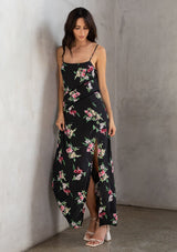 [Color: Black/Red] A brunette model wearing a black dress with a red floral design. A slit maxi dress with a scoop neckline, adjustable spaghetti straps, and lattice trims. A versatile dress for day to night. 