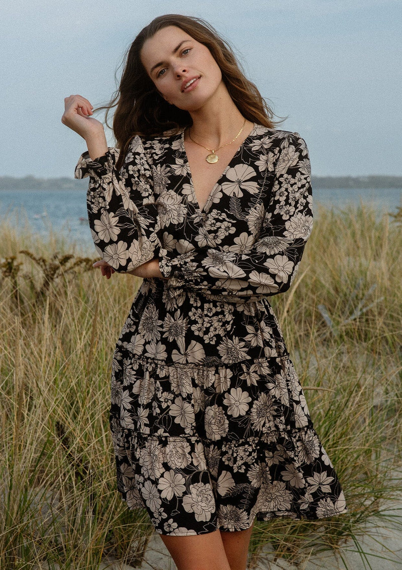 [Color: Black/Taupe] A front-facing image of a brunette model wearing a black and taupe floral print mini dress near the ocean. This casual boho style features a surplice v-neckline, long sleeves with ruffled cuffs, and a flowy tiered skirt. Ideal for casual brunch outings, vacations, and special occasions.