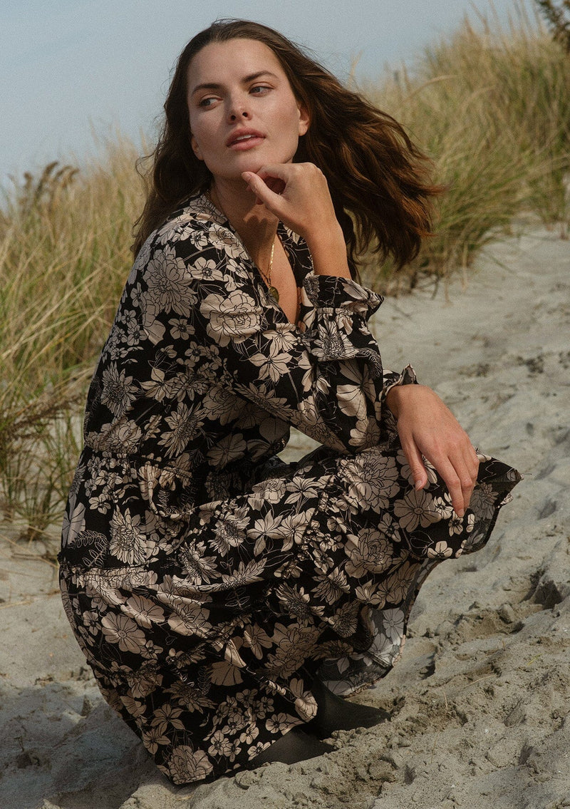 [Color: Black/Taupe] A sitting image of a brunette model wearing a black and taupe floral print mini dress near the ocean. This casual boho style features a surplice v-neckline, long sleeves with ruffled cuffs, and a flowy tiered skirt. Ideal for casual brunch outings, vacations, and special occasions.