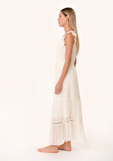 [Color: Vanilla] A side facing image of a blonde model wearing a flowy bohemian white maxi dress with short flutter sleeves, eyelet lace trim, embroidery, and a tassel tie drawstring waist.