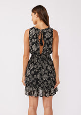 [Color: Black/Natural] A back facing image of a brunette model wearing a sleeveless bohemian mini dress in a black and natural floral print. A chiffon tank dress with a smocked elastic waist and an open back with a tassel tie closure.