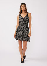 [Color: Black/Natural] A full body front facing image of a brunette model wearing a sleeveless bohemian mini dress in a black and natural floral print. A chiffon tank dress with a smocked elastic waist and an open back with a tassel tie closure.