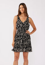 [Color: Black/Natural] A front facing image of a brunette model wearing a sleeveless bohemian mini dress in a black and natural floral print. A chiffon tank dress with a smocked elastic waist and an open back with a tassel tie closure.