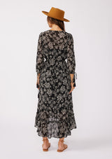 [Color: Black/Natural] A back facing image of a brunette model wearing a bohemian chiffon maxi dress in a black and natural floral print. With a high low ruffled hemline, a button front top, and three quarter length sleeves with tassel tie cuffs.