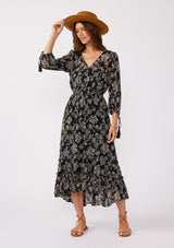 [Color: Black/Natural] A front facing image of a brunette model wearing a bohemian chiffon maxi dress in a black and natural floral print. With a high low ruffled hemline, a button front top, and three quarter length sleeves with tassel tie cuffs.