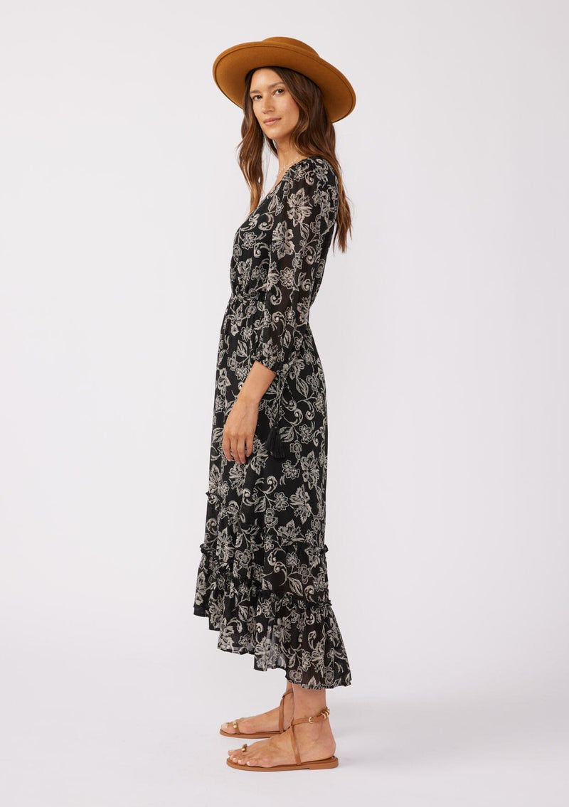 [Color: Black/Natural] A side facing image of a brunette model wearing a bohemian chiffon maxi dress in a black and natural floral print. With a high low ruffled hemline, a button front top, and three quarter length sleeves with tassel tie cuffs.