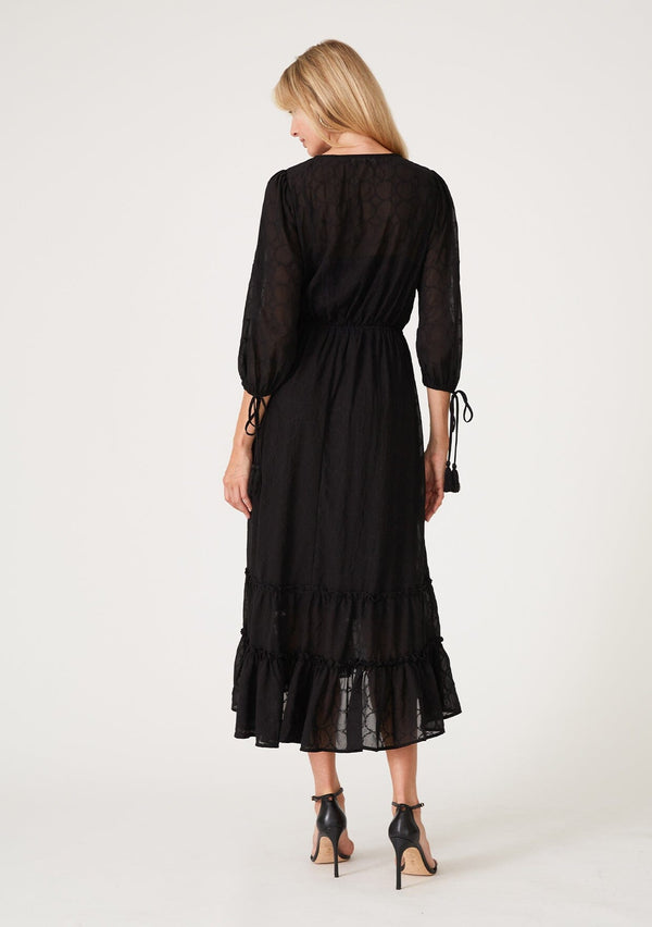 [Color: Black] A back facing image of a blonde model wearing a bohemian black maxi dress in embroidered chiffon. With three quarter length sleeves, adjustable tie cuffs, a self covered button front, an elastic waist, and a ruffle trimmed tiered skirt.