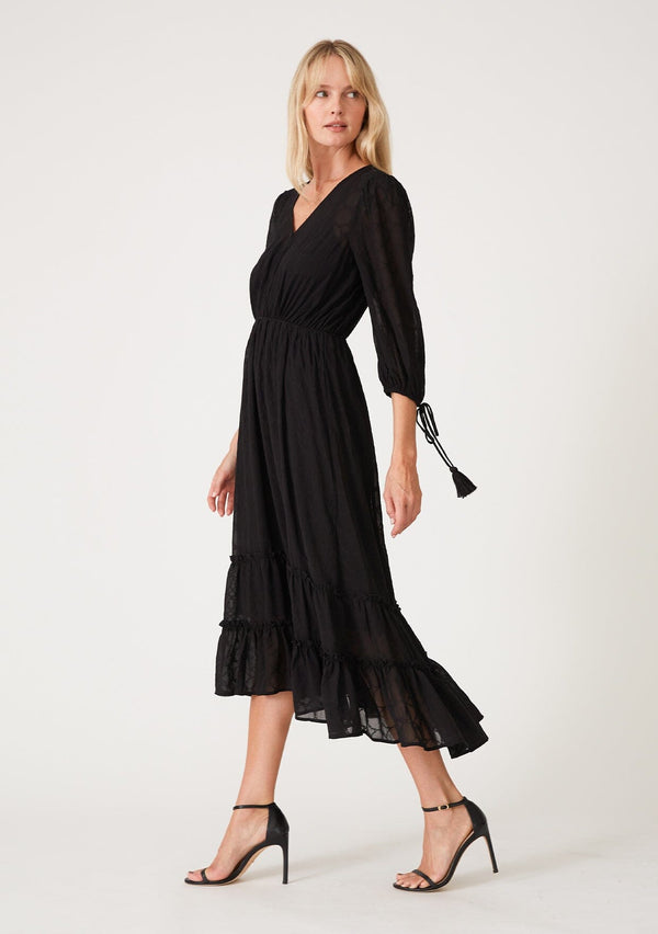 [Color: Black] A side facing image of a blonde model wearing a bohemian black maxi dress in embroidered chiffon. With three quarter length sleeves, adjustable tie cuffs, a self covered button front, an elastic waist, and a ruffle trimmed tiered skirt.