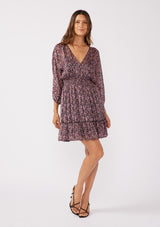 [Color: Black/Plum] A full body image of a brunette model wearing a purple floral print mini dress with shimmering metallic stripes. This holiday dress features a flattering v-neckline, 3/4 length sleeves, a smocked waist, and a ruffle-trimmed skirt. Ideal for special occasions and festive events.