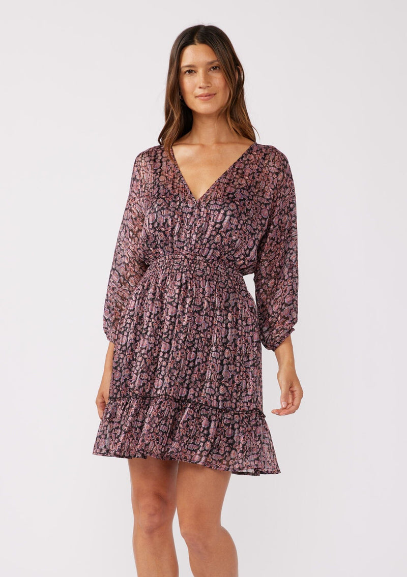 [Color: Black/Plum] A front facing image of a brunette model wearing a purple floral print mini dress with shimmering metallic stripes. This holiday dress features a flattering v-neckline, 3/4 length sleeves, a smocked waist, and a ruffle-trimmed skirt. Ideal for special occasions and festive events.