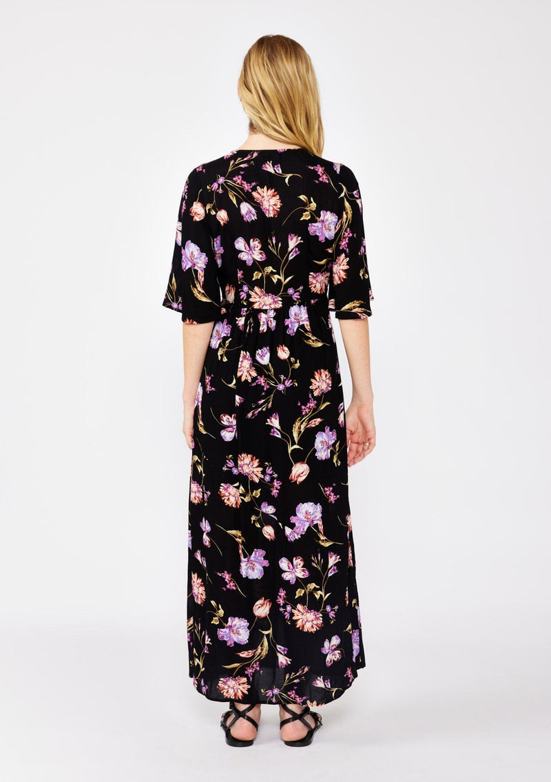 [Color: Black/Purple] A blonde model wearing a classic bohemian silhouette in a black and purple floral print. A relaxed fit maxi dress with flowy short sleeves, plunging v neckline, empire waistline with a front elastic waist, and side slit. Styled with casual black strappy sandals for a day to night look.