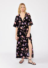 [Color: Black/Purple] A blonde model wearing a classic bohemian silhouette in a black and purple floral print. A relaxed fit maxi dress with flowy short sleeves, plunging v neckline, empire waistline with a front elastic waist, and side slit. Styled with casual black strappy sandals for a day to night look.