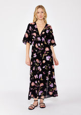 [Color: Black/Purple] A blonde model wearing a classic bohemian silhouette in a black and purple floral print. A relaxed fit maxi dress with flowy short sleeves, plunging v neckline, empire waistline with a front elastic waist, and side slit. Styled with casual black strappy sandals for a day to night look.